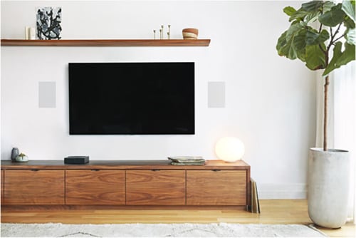 Sonos In-Wall Speaker model INWLLWW1 installed on each side of a mounted TV in a modern living room with wood furniture, white walls, and a tall potted plant.