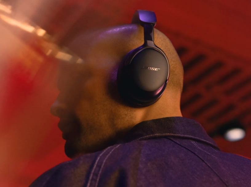 Person wearing black Bose QuietComfort Ultra Headphones 2nd Gen with gold logo branding, shown from behind against a warm red-toned background with motion blur effects. view