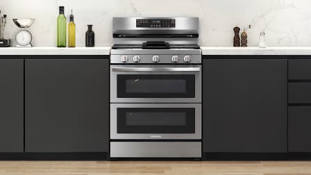 Front view of Samsung NX60A6751SS stainless steel gas double oven range with five burner cooktop, installed in kitchen with black cabinets and white countertops.