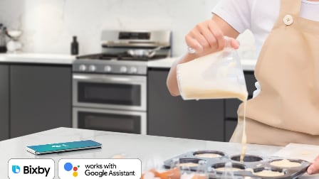 Person pouring batter into muffin tin with Samsung double oven range in background, Bixby and Google Assistant logos showing Wi-Fi and voice control compatibility