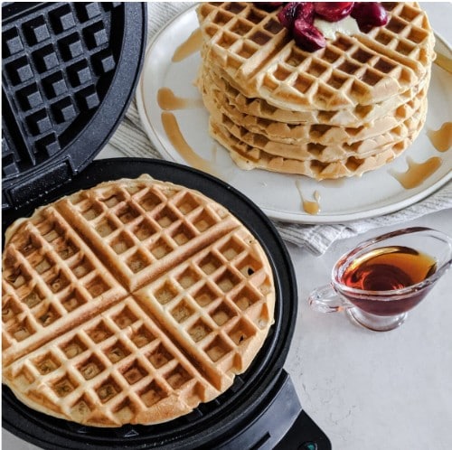 Cuisinart WMRCA waffle maker open with a golden waffle inside, next to a stack of waffles topped with fruit and syrup on a white plate, with a small pitcher of syrup beside it.