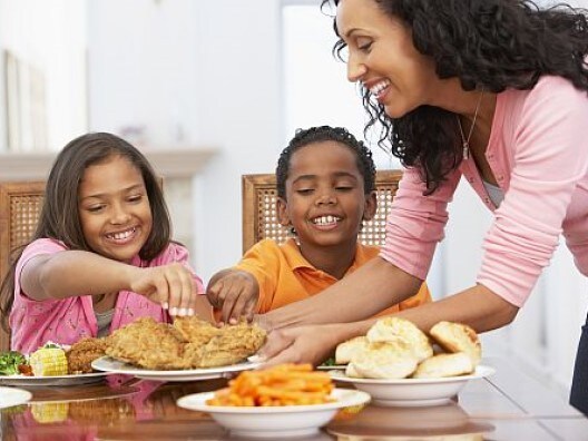Two kids sitting at table taking fried chicken from a dish their mother is setting down