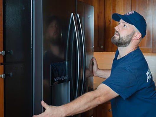 A man in a hat holding the sides of a refrigerator to put it in place in a kitchen.
