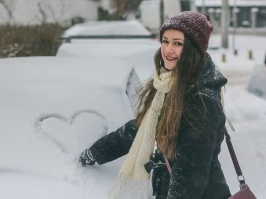 A smiling woman drawing a heart on the snowy windshield of a car